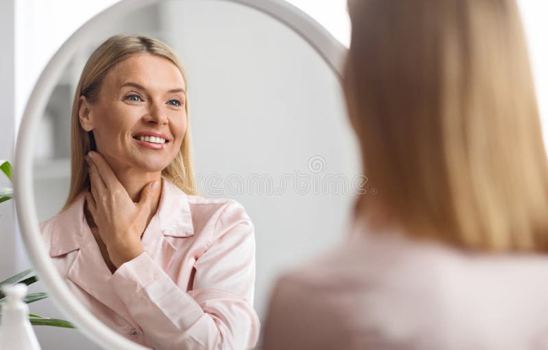hermosa-mujer-de-mediana-edad-mirando-al-espejo-y-tocándose-la-piel-del-cuello-425968159